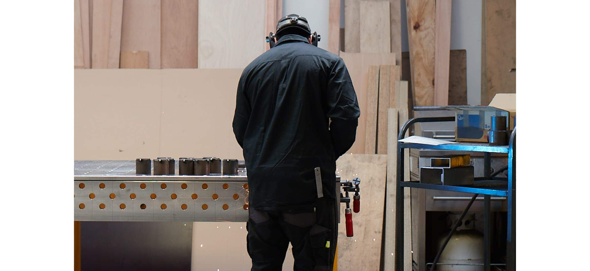 A metal worker at a bench in a workshop with their back to the camera. 